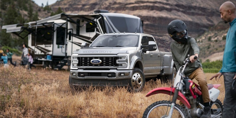 An all-new Silver 2024 Ford F-350 parked in a field with a fifth wheel and dirt bike. - Jack Madden Ford Norwood, MA