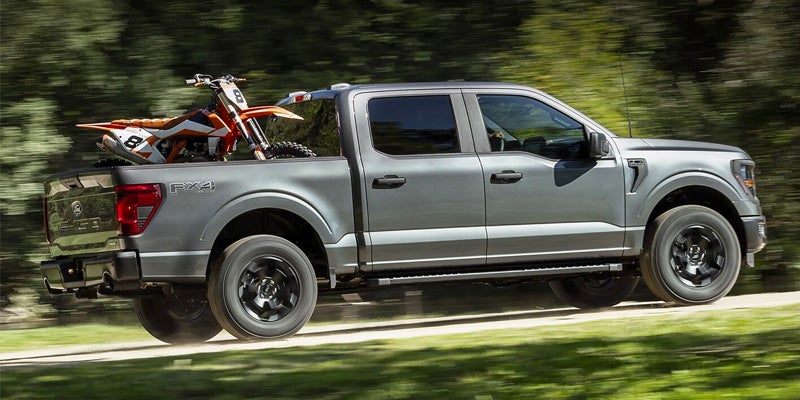 An all-new Gray 2024 Ford F-150 hauling an orange dirt bike up a tree covered hill. - Jack Madden Ford Norwood, MA