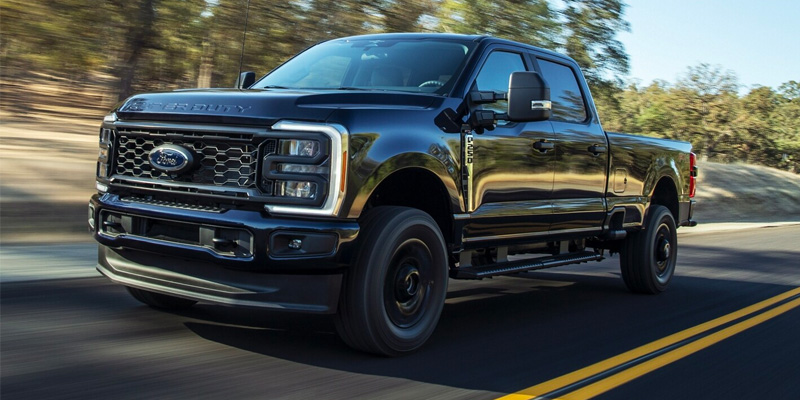 An all-new black Ford Super Duty driving down a tree-lined highway road. - Jack Madden Ford Norwood, MA