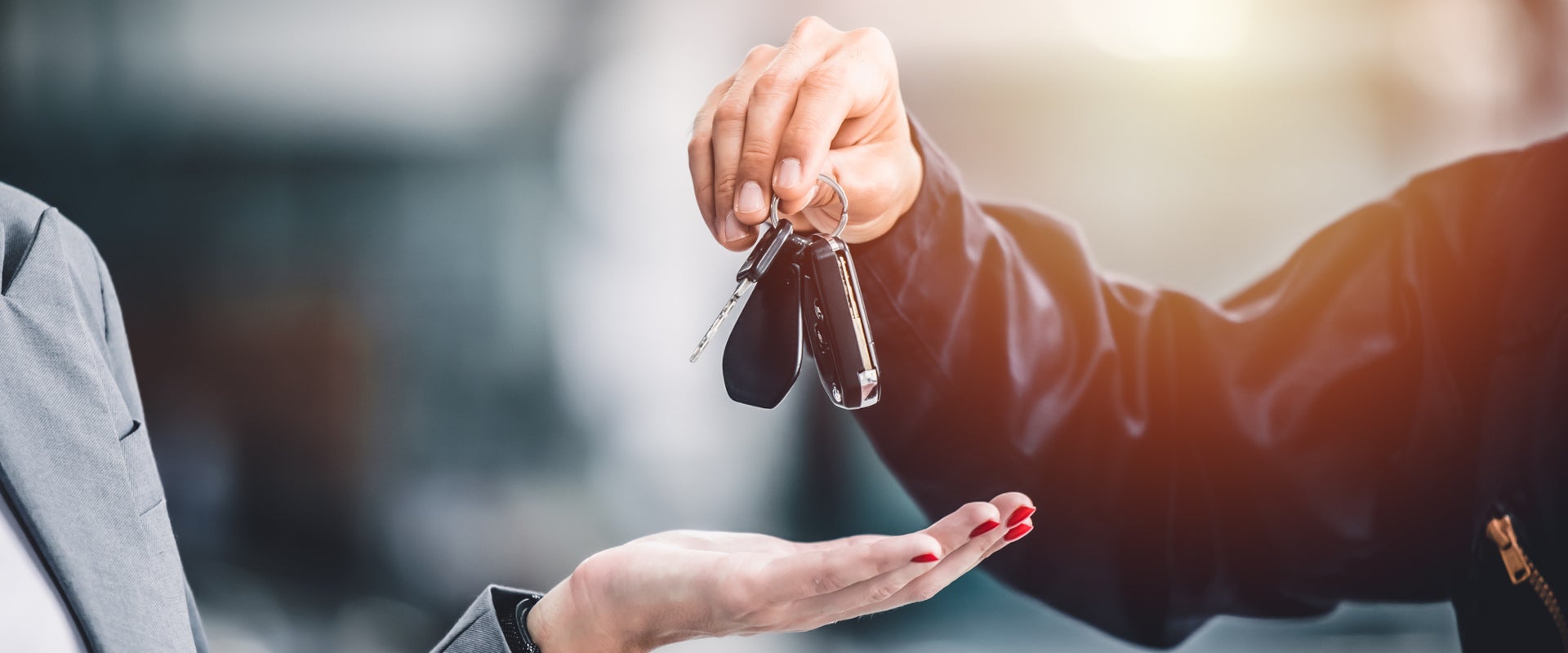 A close-up of two hands exchanging car keys, one hand with red nails receiving from another in a dark sleeve. The scene conveys a sense of trust and transaction.