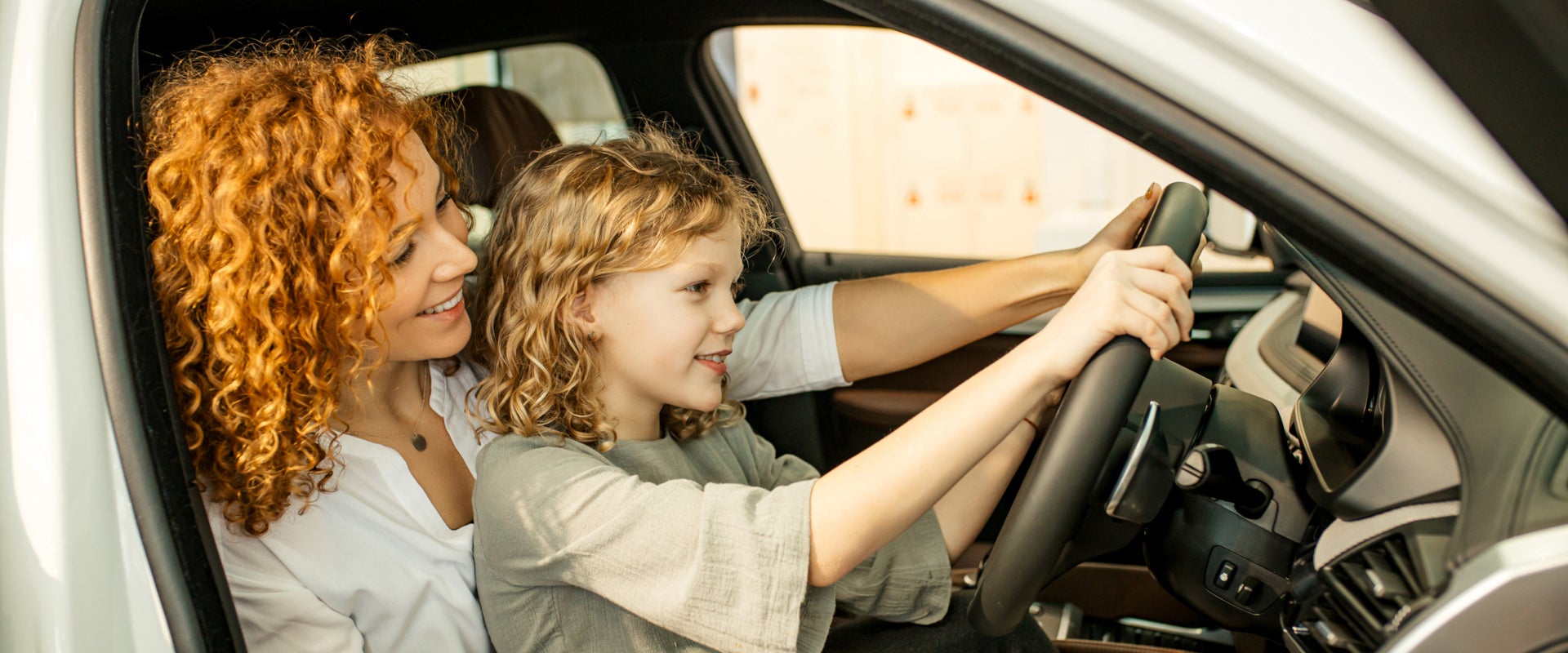 A curly-haired woman and a young girl share a joyful moment in a car. The girl holds the steering wheel, smiling, while the woman watches with affection.